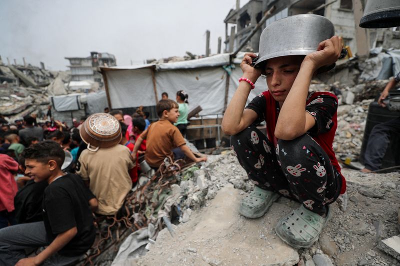 FILE PHOTO: A girl puts a pot to her head as Palestinians wait to receive food cooked by a charity kitchen, in Beit Lahia, northern Gaza Strip, April 24, 2025. REUTERS/Mahmoud Issa/File Photo